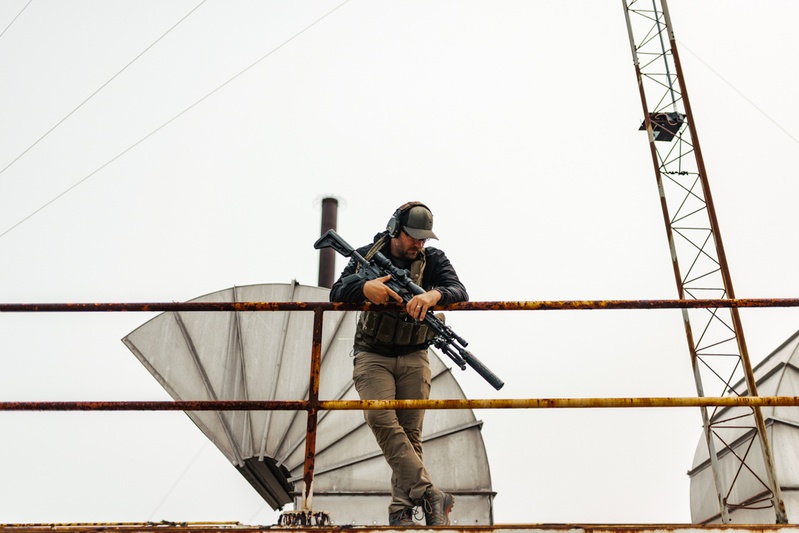 Jared standing on a rusted steel catwalk at a long-range course, holding a rifle and wearing ear protection