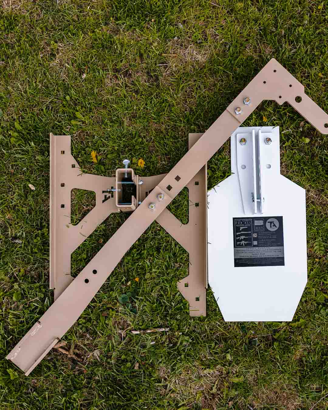 Steel target parts on grass: tan steel base frame, 2x4 upright bracket, and white C-zone steel target plate